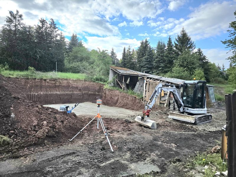 Frisch ausgehobene Baugrube mit Vermessungsstativ und Yanmar-Bagger